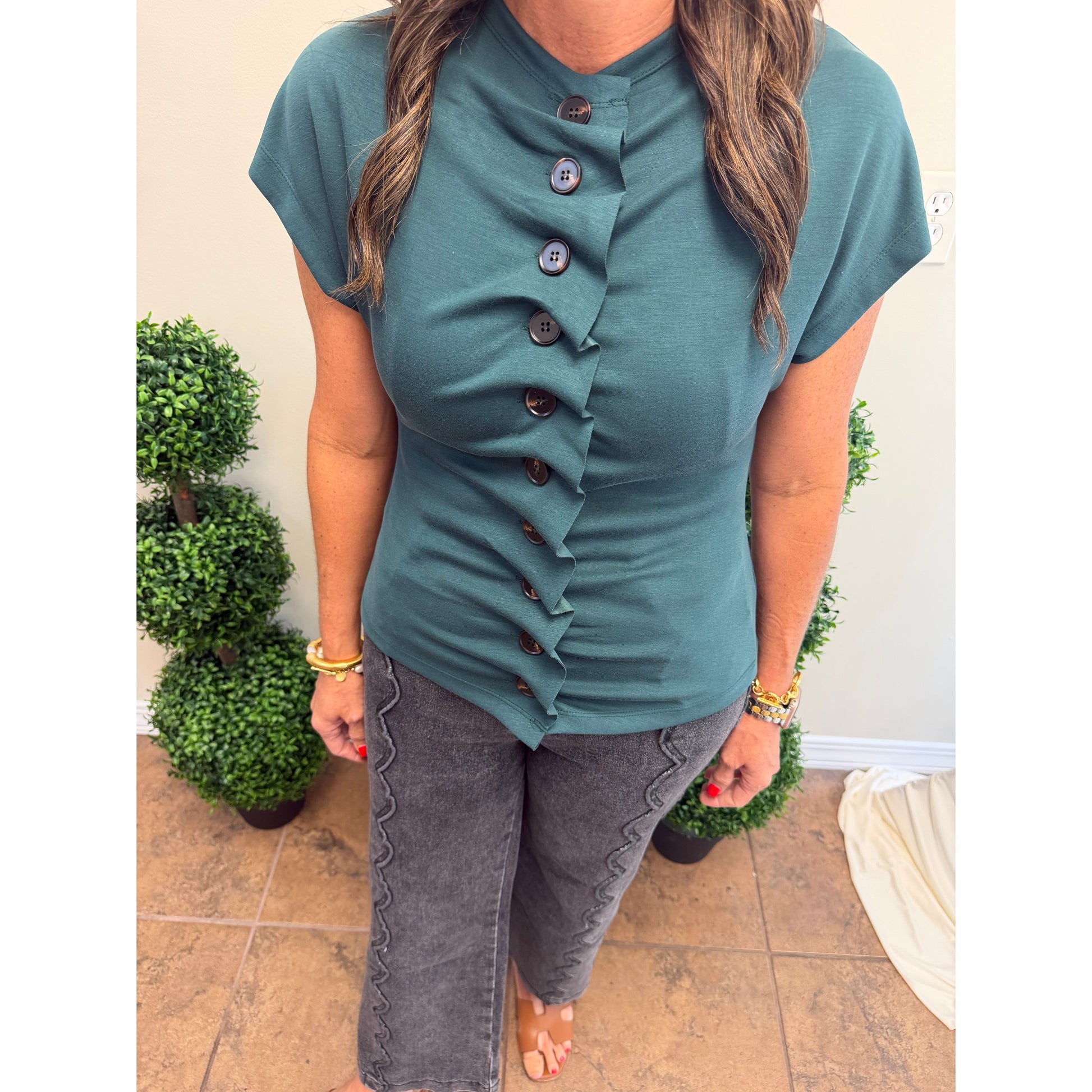 A woman wearing a teal short-sleeve top with a ruffled button-front detail and dark gray scalloped pants, standing indoors between potted plants.