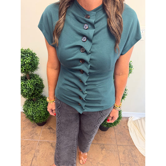 A woman wearing a teal short-sleeve top with a ruffled button-front detail and dark gray scalloped pants, standing indoors between potted plants.