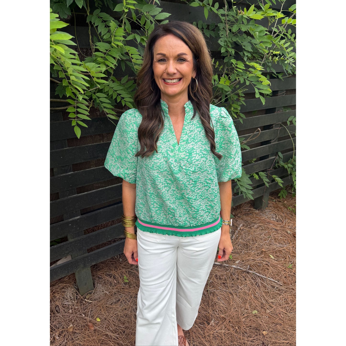 Woman in a green floral top and white pants standing outdoors with plants and ground visible.