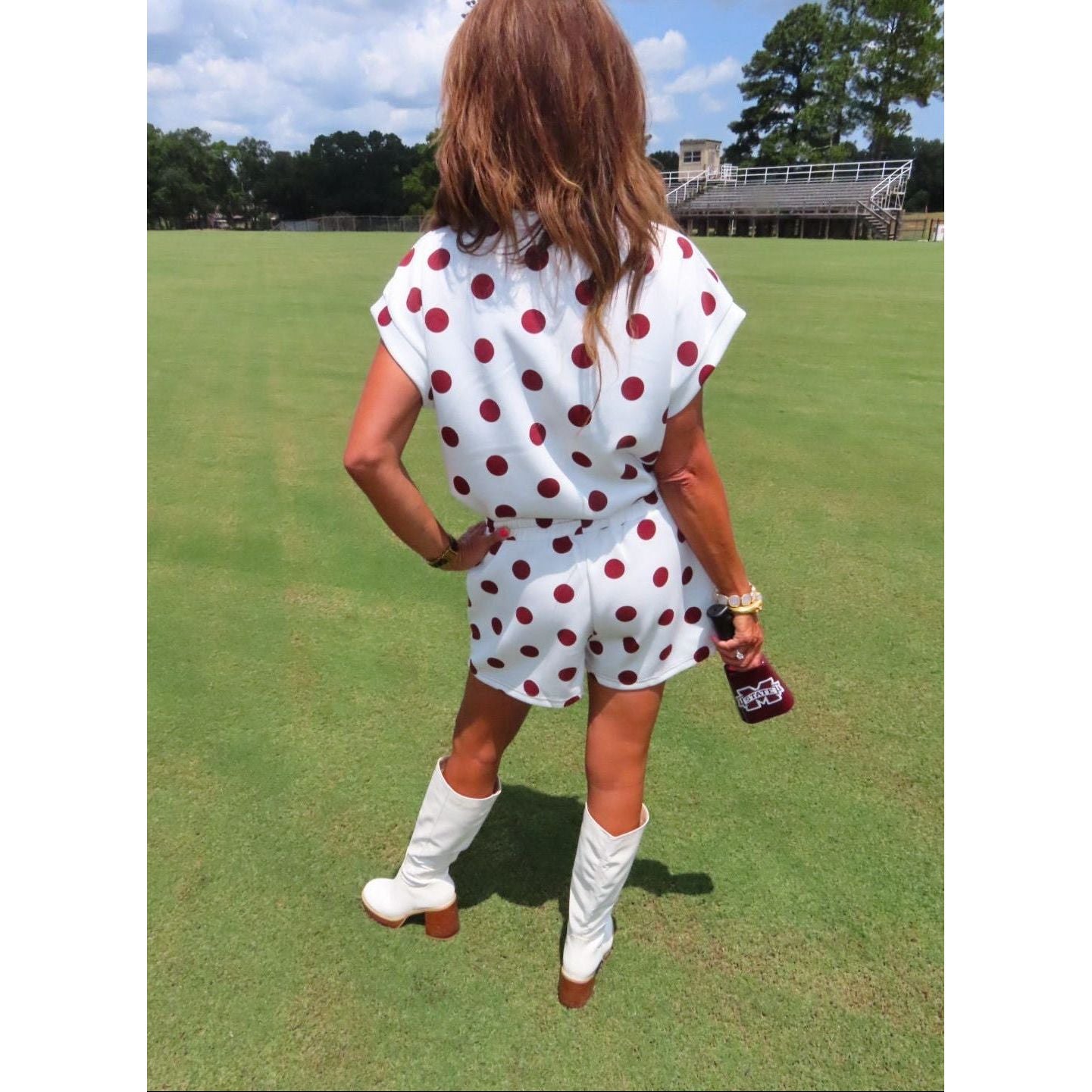 A woman stands on a grassy field wearing a white short-sleeve romper with red polka dots, white knee-high boots, and holding a brown bottle.