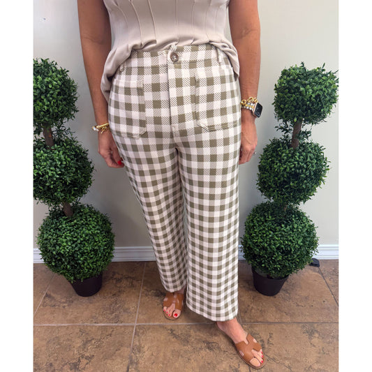 A person wearing high-waisted, cropped wide-leg trousers in a beige and brown gingham check pattern, styled with a beige top and tan slide sandals, standing in front of a wall with decorative potted topiary trees.
