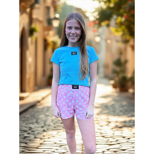 Young girl wearing a blue t-shirt and pink shorts with white polka dots, standing on a stone path.