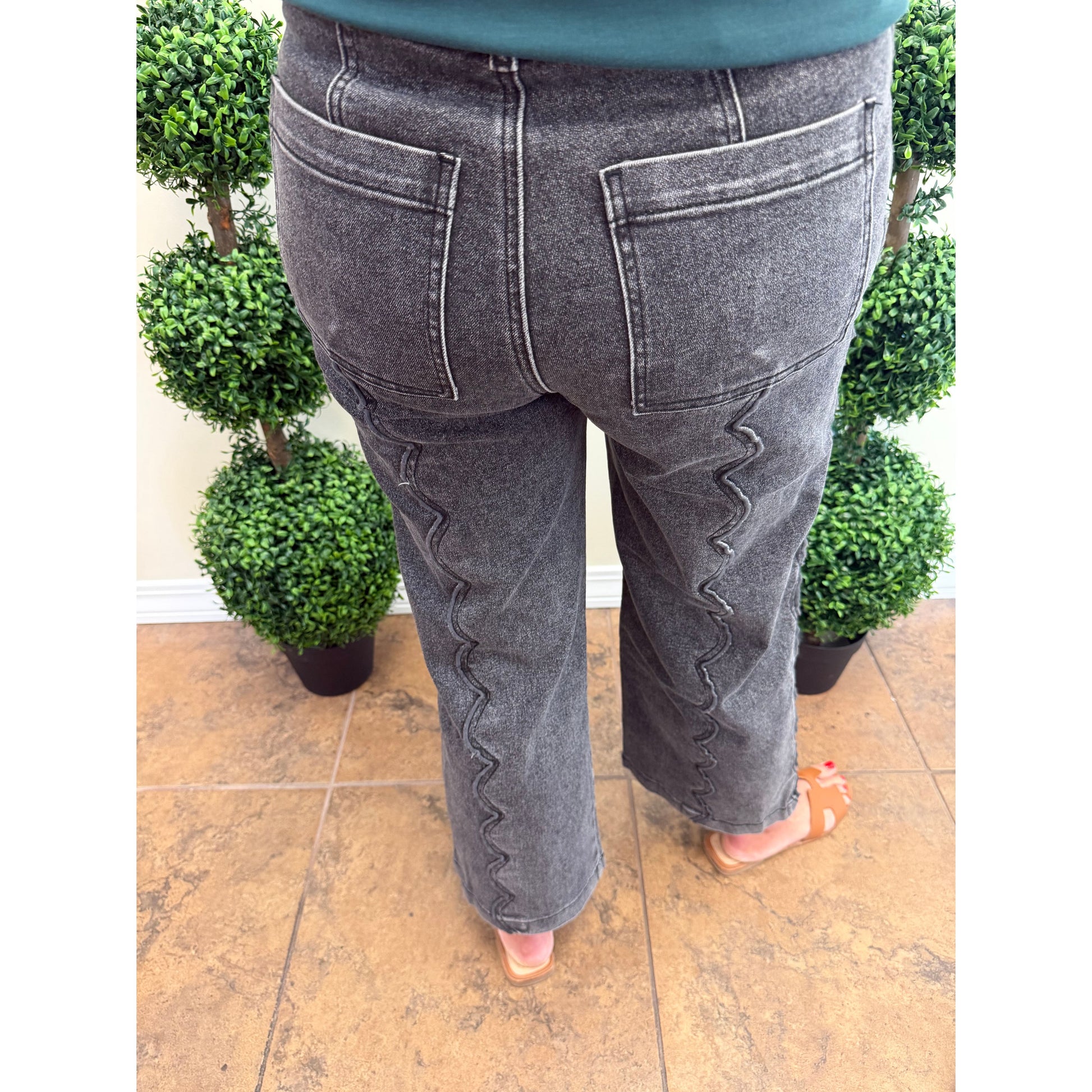 A person viewed from behind wearing dark gray denim jeans with distinctive zigzag contrast stitching down the back of both legs, standing on a tiled floor between potted green plants.