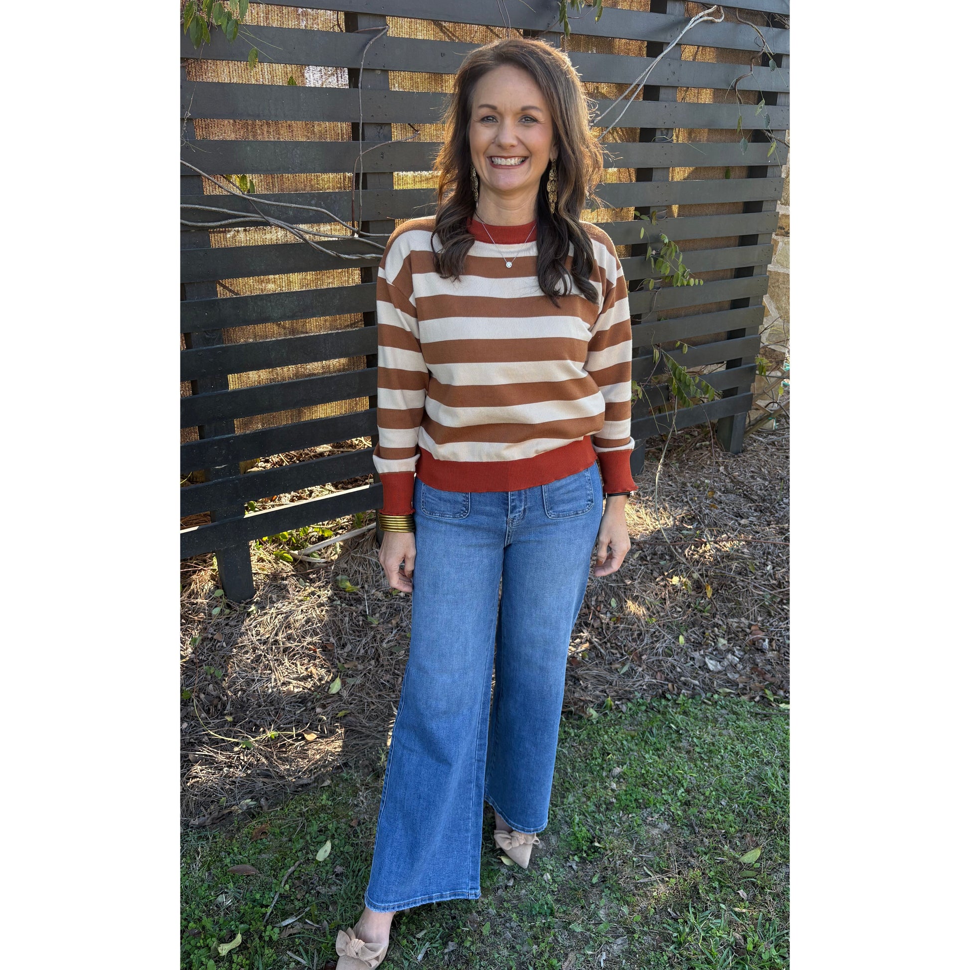 Person wearing a striped sweater and blue jeans standing outdoors near a wooden fence.