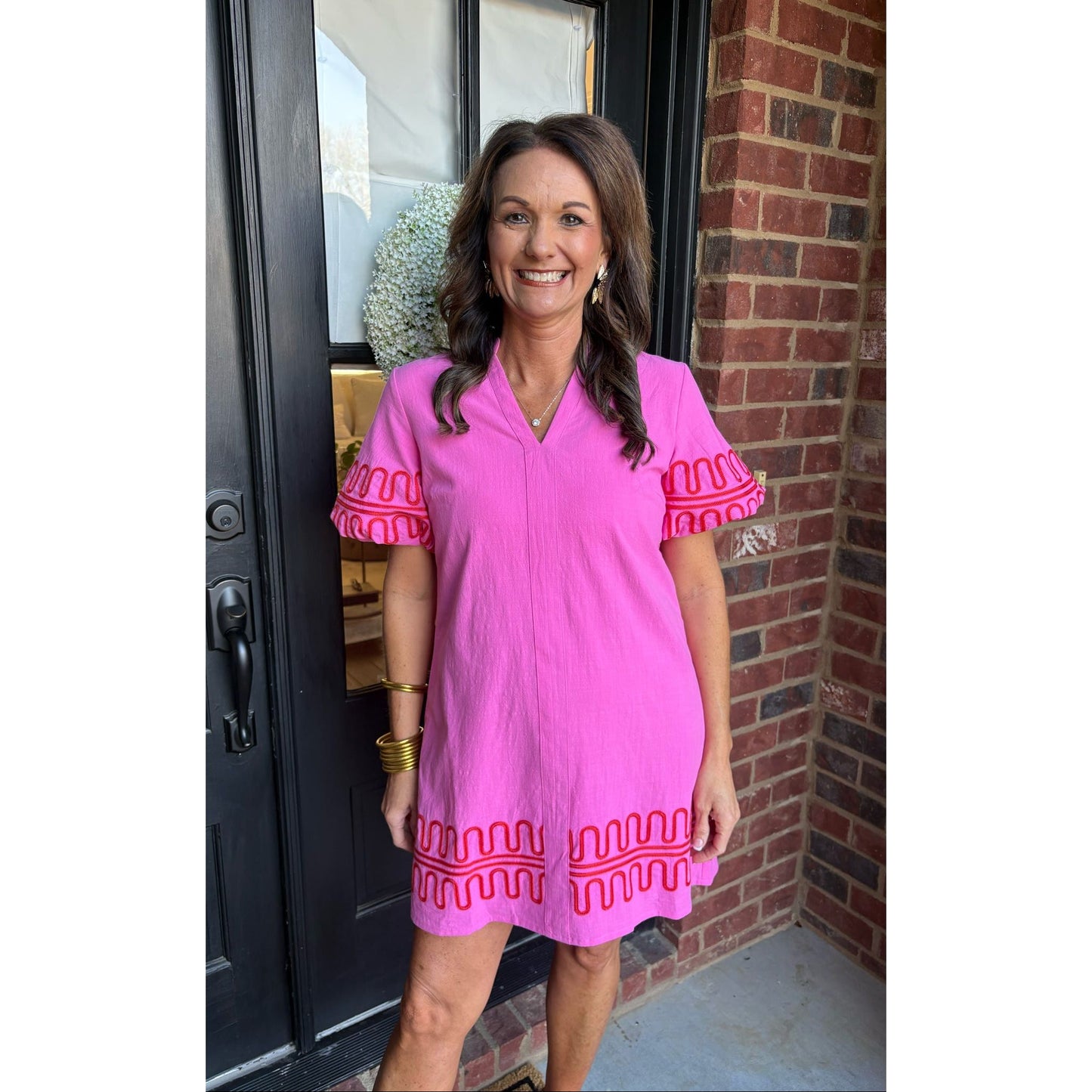 Woman wearing a pink dress with red embroidery standing in front of a brick wall.