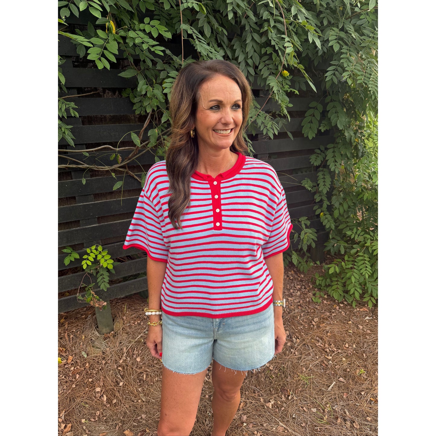 A woman wearing a red and white striped short-sleeve henley top with light blue denim shorts, standing outdoors in front of a dark wooden fence and green foliage.
