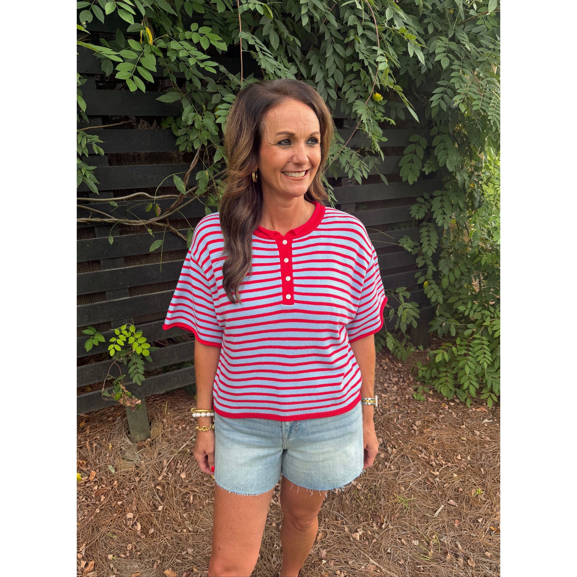 A woman wearing a red and white striped short-sleeve henley top with light blue denim shorts, standing outdoors in front of a dark wooden fence and green foliage.