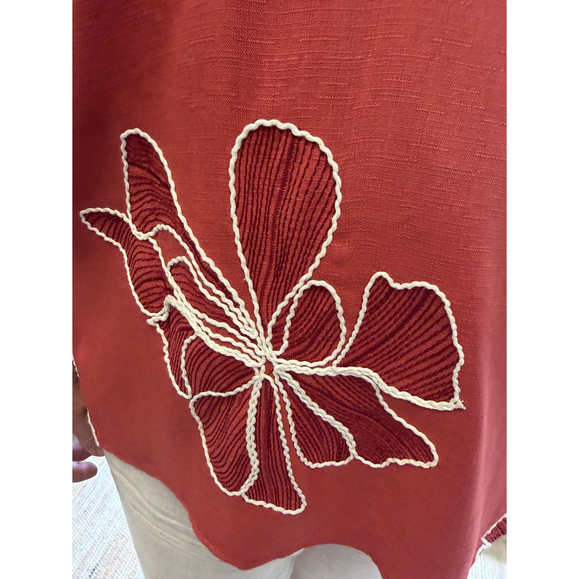 A close-up of a terracotta red linen garment featuring a large, raised cream-colored floral embroidery on the front.
