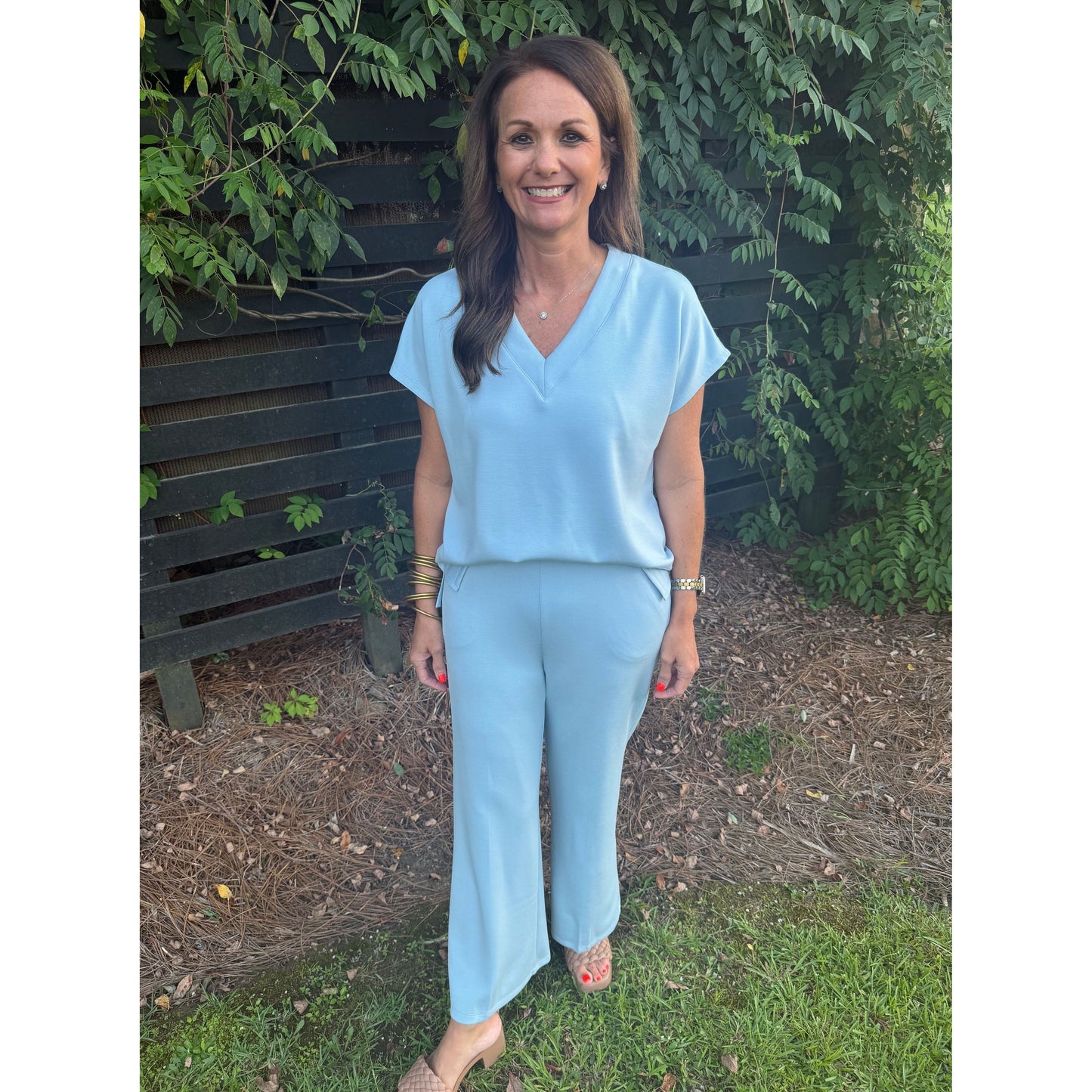 A woman wearing a light blue matching two-piece set consisting of a short-sleeve V-neck top and wide-leg pants, standing outdoors in a garden setting with green foliage and a dark wooden fence behind her.