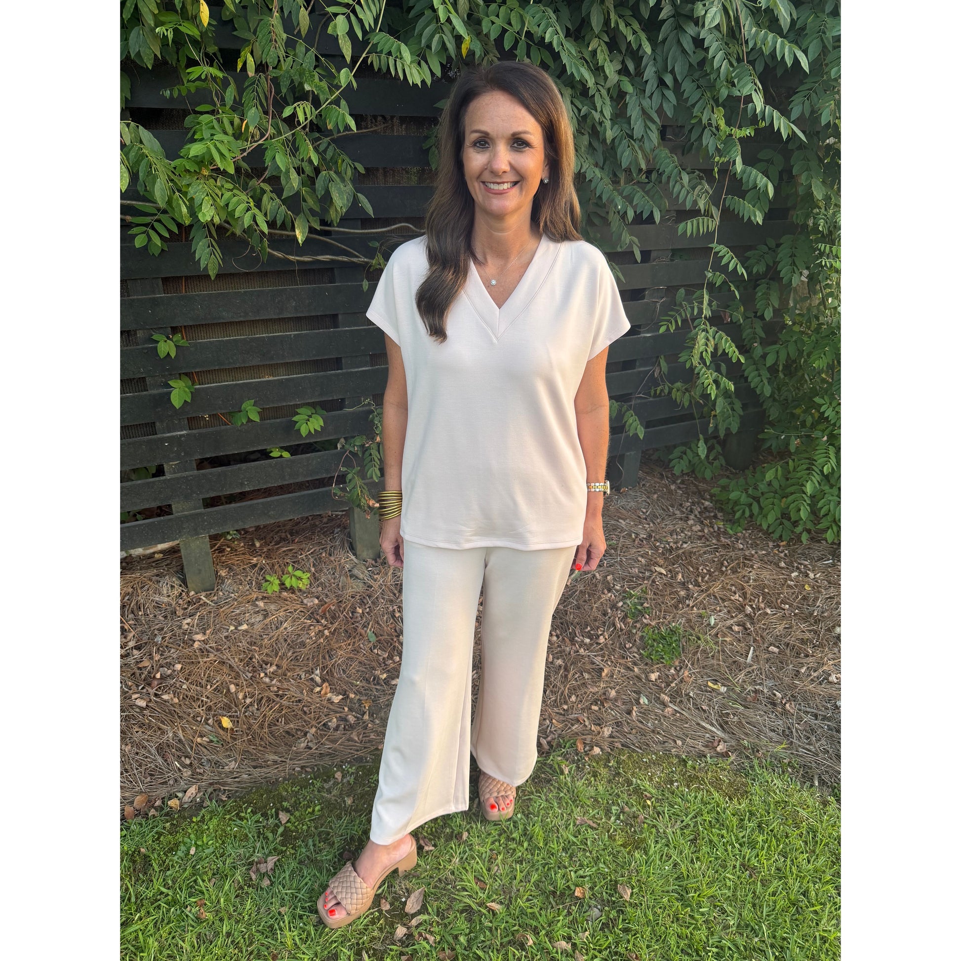 A woman wearing a matching cream-colored short-sleeve V-neck top and wide-leg pants stands outdoors on a grassy lawn in front of a dark wooden fence and green foliage.