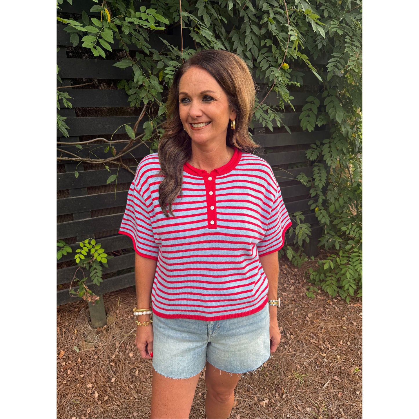 A woman wearing a red and white striped short-sleeve henley top with light blue denim shorts, standing outdoors in front of a green leafy fence.