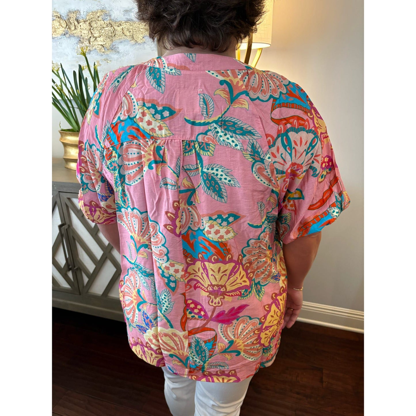 Person wearing a colorful pink patterned shirt in a room with a plant and lamp.