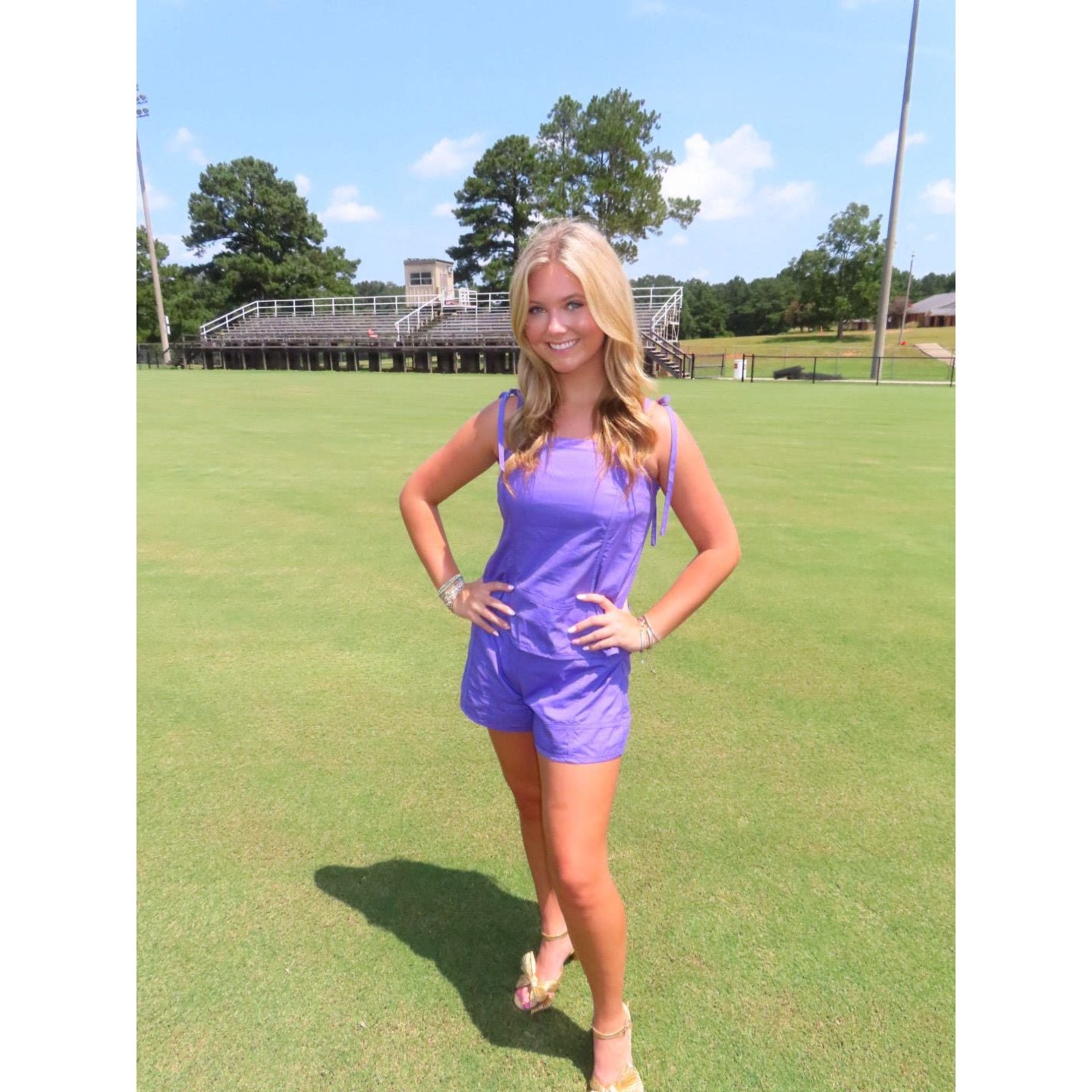 A woman posing on a green grass field wearing a shiny purple sleeveless romper and beige sandals, with bleachers and trees in the background.