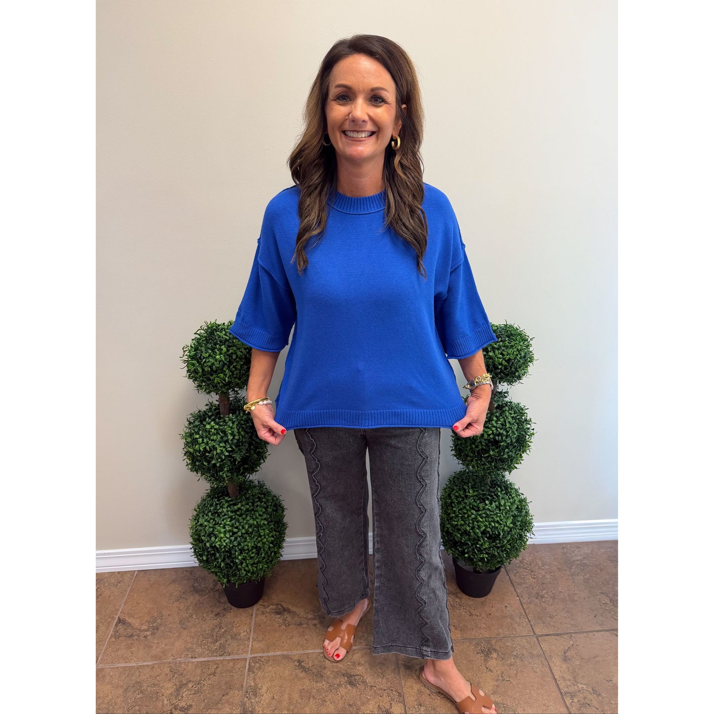 A woman modeling a bright blue short-sleeve knit sweater and dark gray textured wide-leg pants, standing indoors between two potted green topiary trees against a plain beige wall.