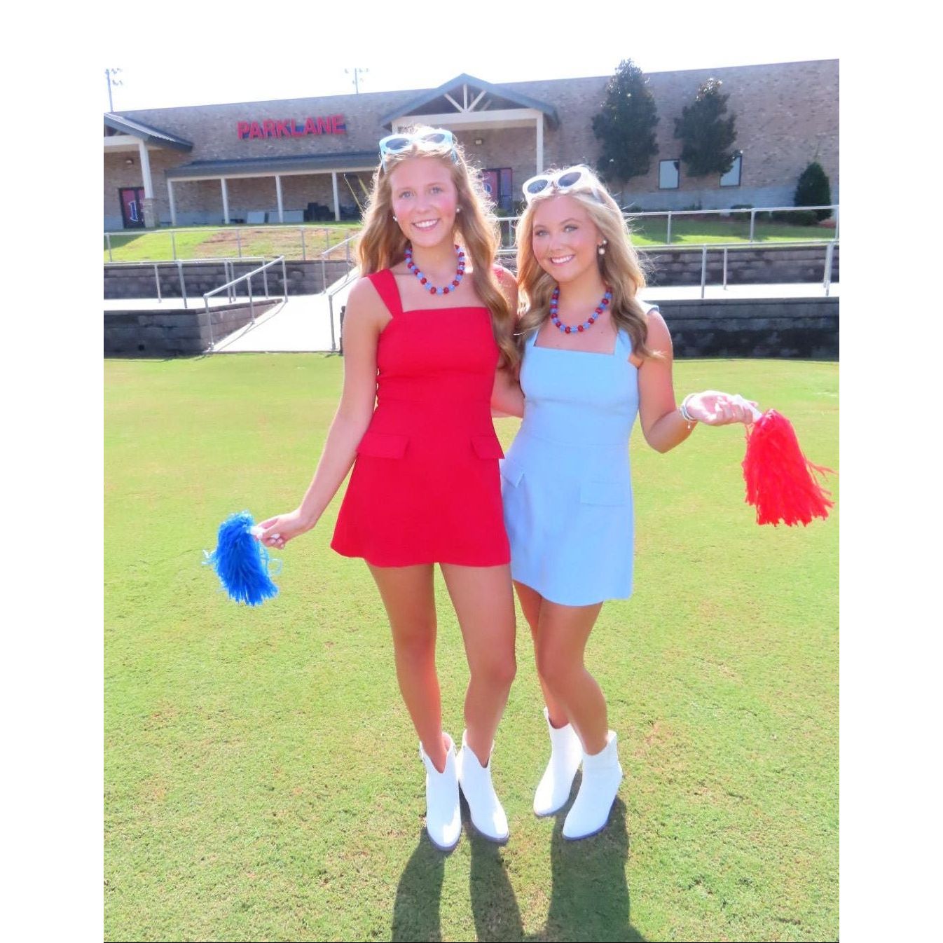 Two women standing on a grassy field in front of a building, wearing bright red and light blue mini dresses with white ankle boots and holding blue and red pom-poms.