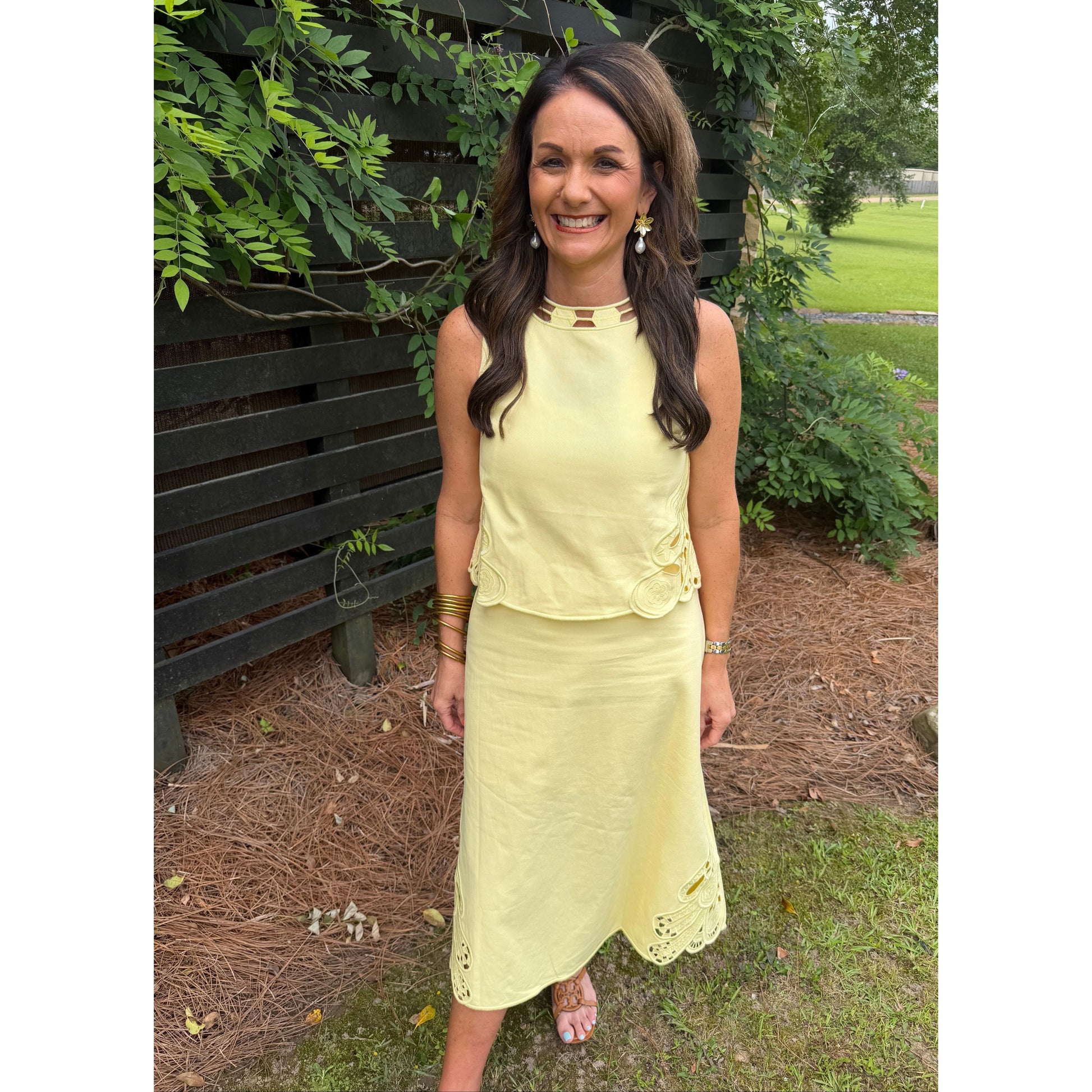 Woman in a yellow dress standing outdoors on a bench with greenery around.