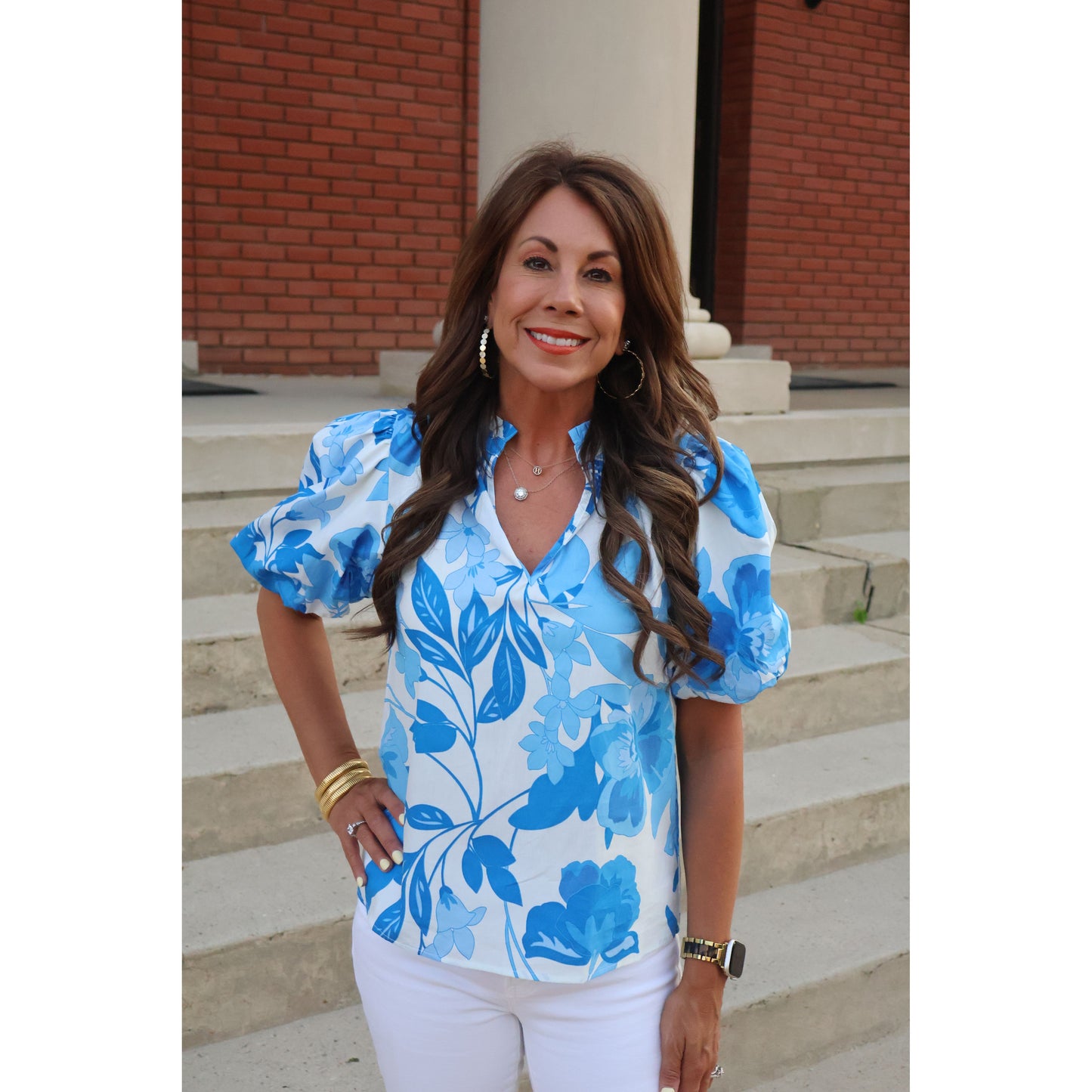 woman standing on steps wearing blue and white floral top with white pants. 