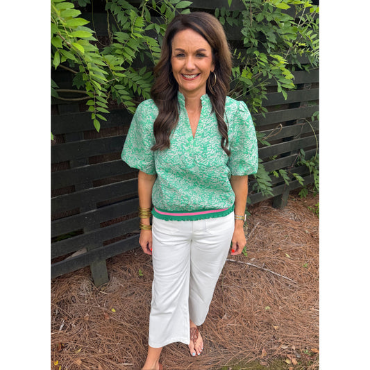 Woman in a green patterned top and white pants standing outdoors with greenery around.