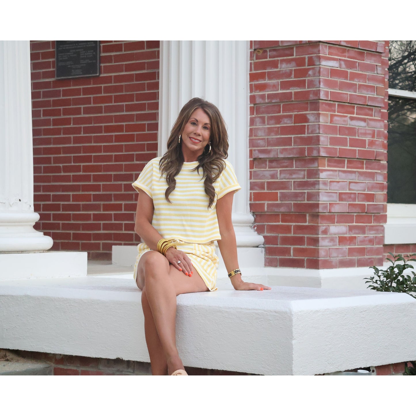 Woman in a yellow outfit sitting on a white bench in front of a brick building.