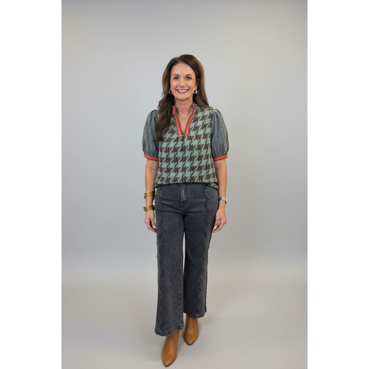 A woman stands indoors wearing a patterned short-sleeve top with puff sleeves and dark gray cropped jeans. The top has a brown and gray houndstooth-style pattern with muted green and blue tones, and red-orange trim at the neckline and sleeve edges.