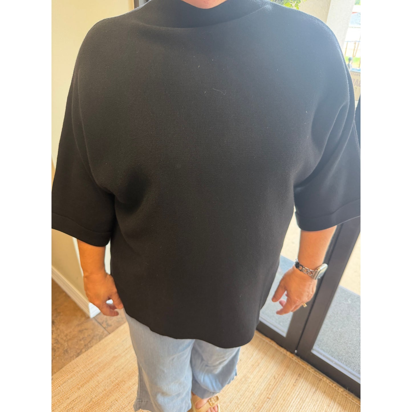A person wearing an oversized black short-sleeve t-shirt and light blue jeans, standing indoors near a glass door.
