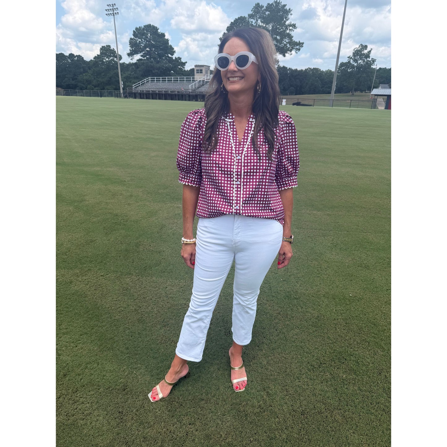A woman stands on a grassy field wearing a pink and white gingham button-up blouse, white pants, and white sandals.