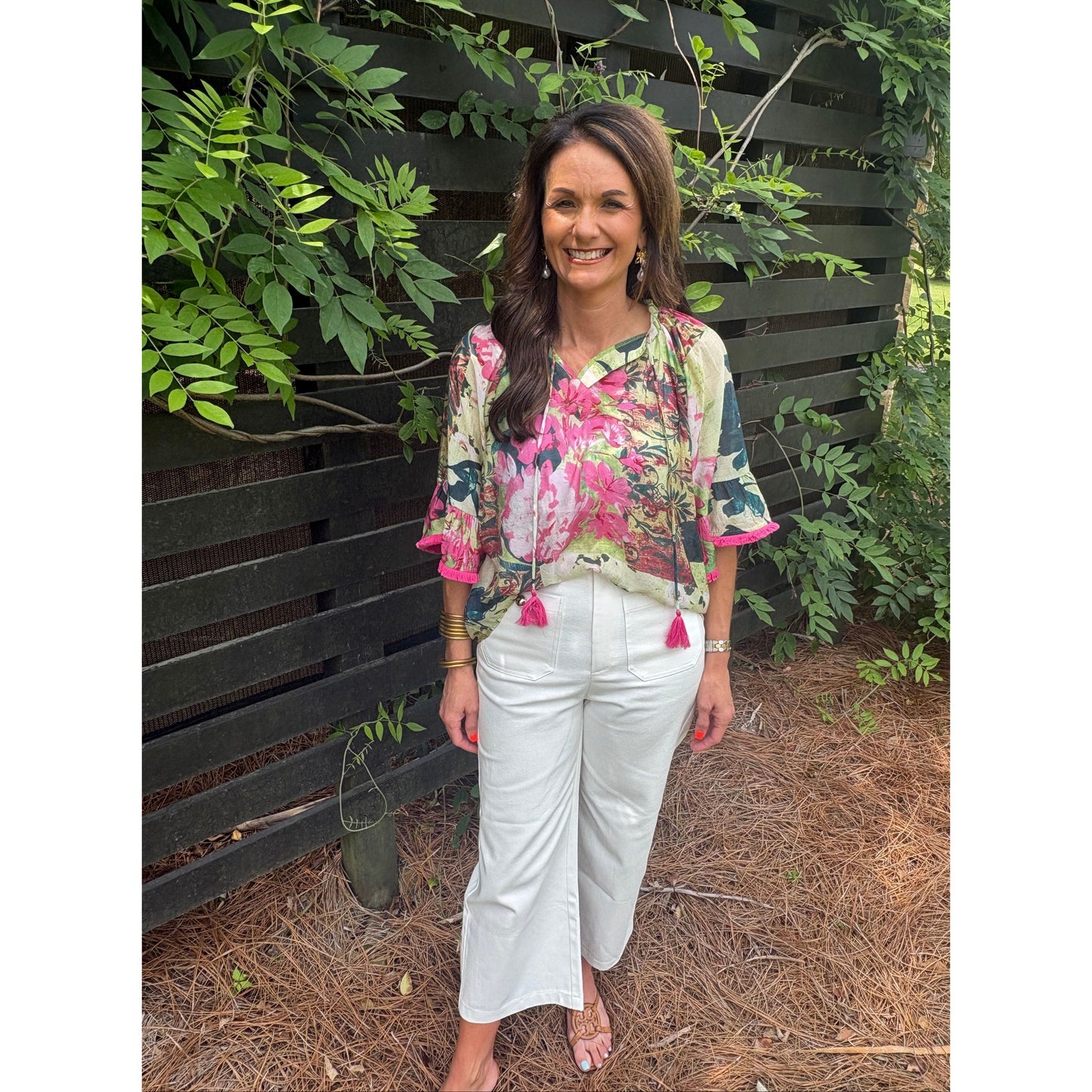 Woman in a floral top and white pants standing outdoors with greenery around.