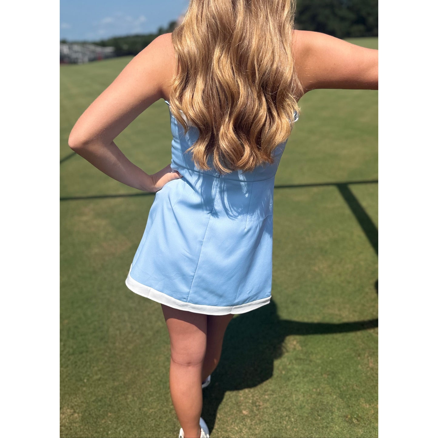A woman standing on a grassy field wearing a light blue sleeveless mini dress with white trim, photographed from behind.