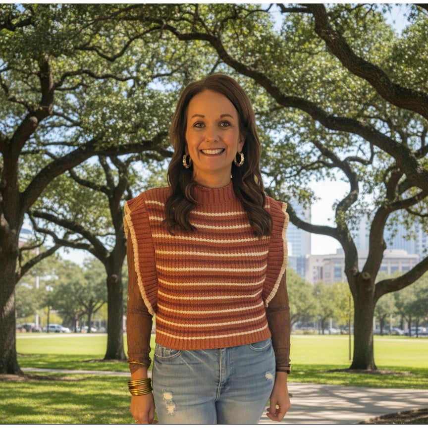 Woman wearing a striped sweater and jeans standing in front of a window with blinds.