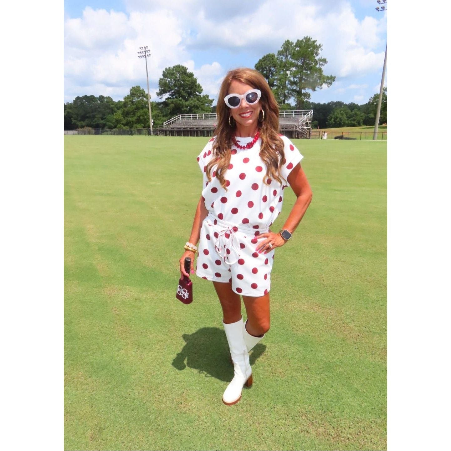 A woman stands on a green grass field wearing a white short-sleeve romper with red polka dots, white knee-high boots, and white sunglasses. She is holding a brown bottle, with trees, stadium lights, and a cloudy sky in the background.