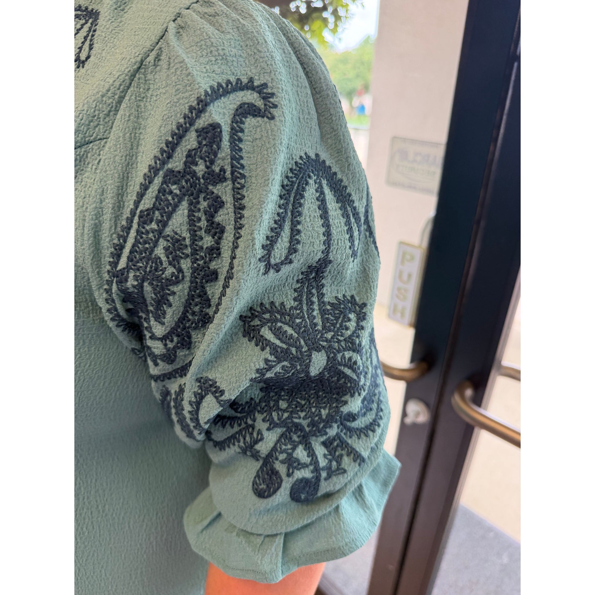 A close-up of a person wearing a sage green short-sleeve blouse with dark blue-gray embroidered paisley patterns on the sleeve, standing near a doorway.
