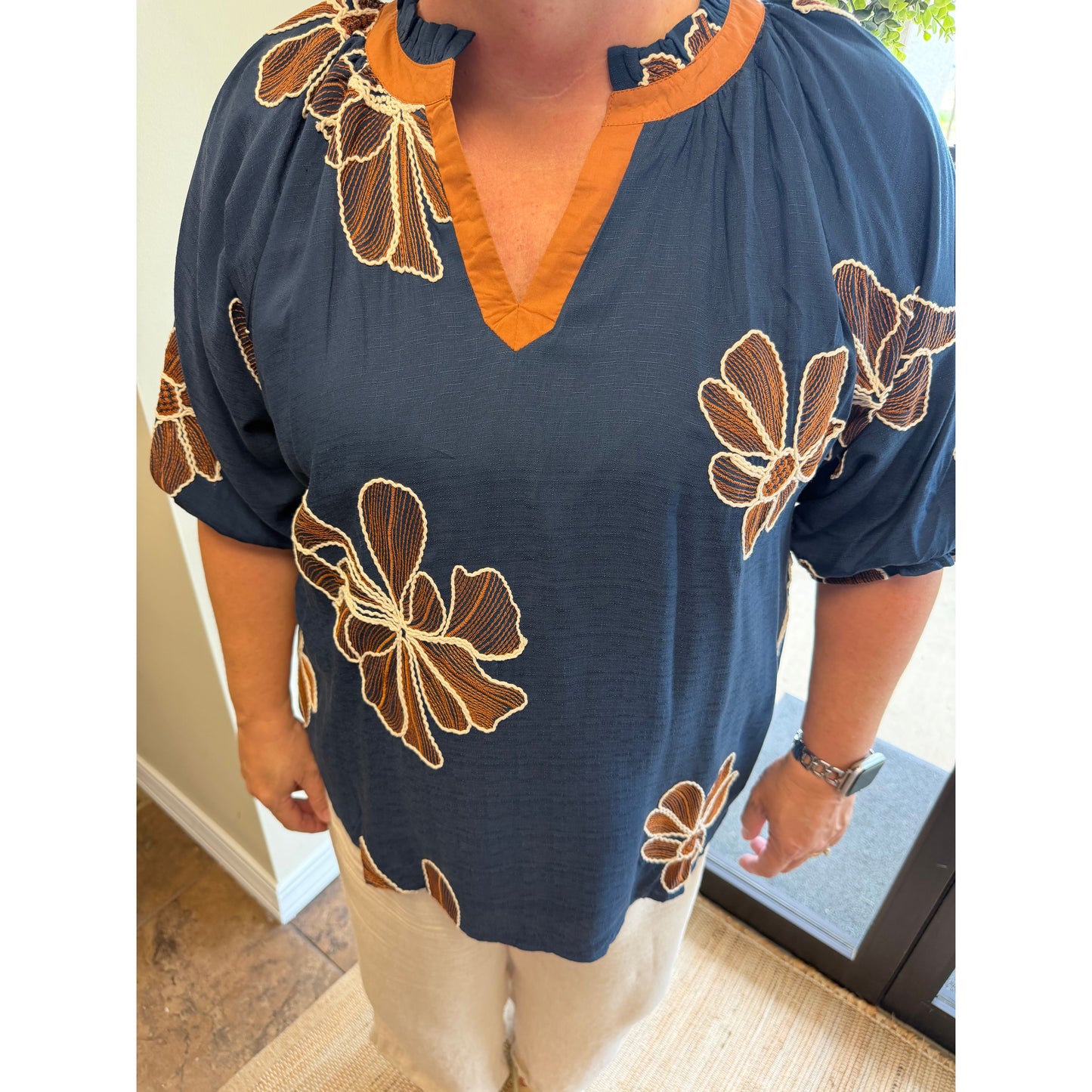 A woman wearing a navy blue blouse with brown and beige floral embroidery and a contrasting orange V-neck trim, paired with beige pants.