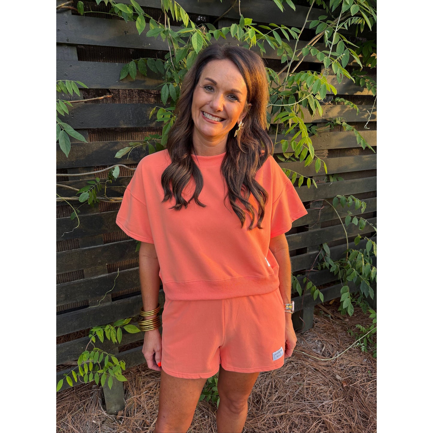Woman wearing a coral outfit standing in front of a wooden fence with greenery.
