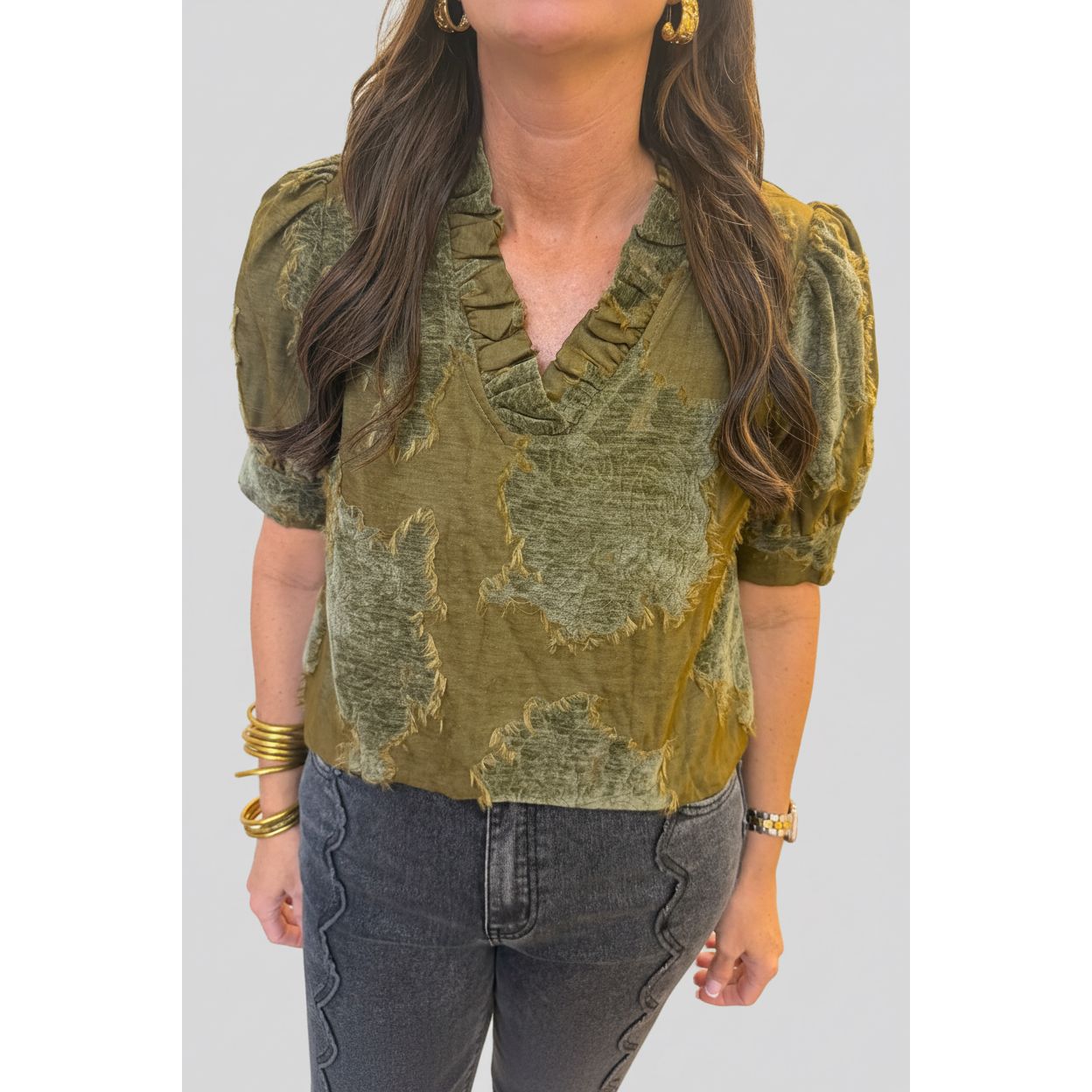 A woman wearing a distressed olive green short-sleeve blouse with puff sleeves and a ruffled V-neckline, paired with dark gray jeans.