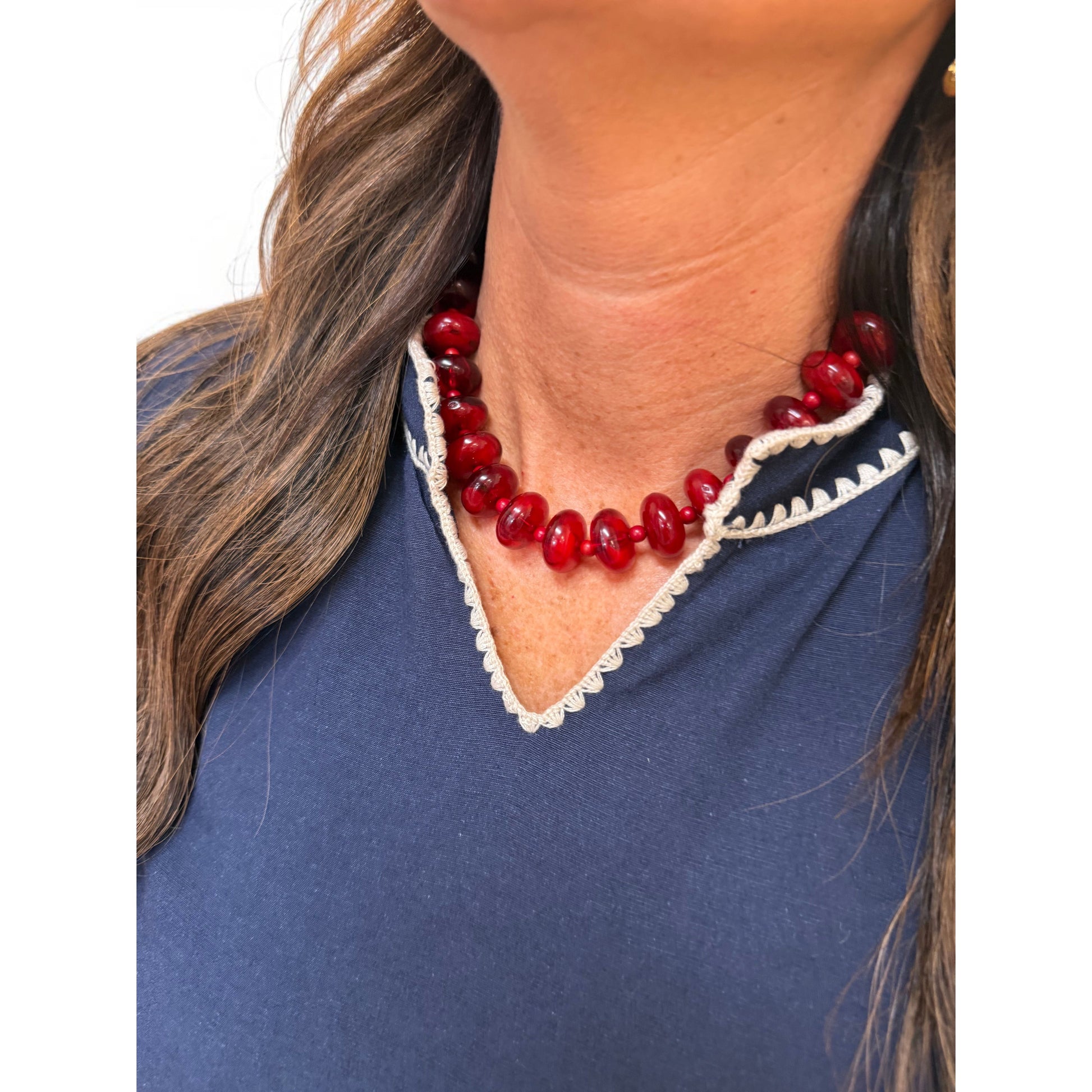 A close-up of a woman wearing a chunky red beaded necklace with a white scalloped trim, paired with a navy blue top.