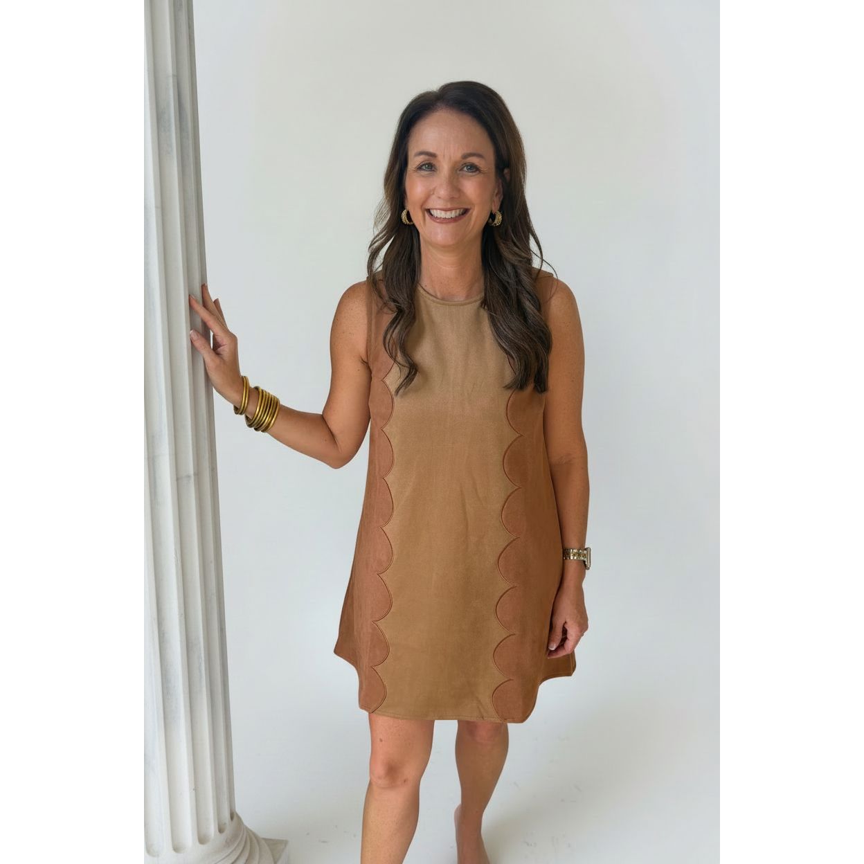 A woman wearing a tan sleeveless dress with scalloped side panels, standing on a covered outdoor walkway next to a beige pillar.