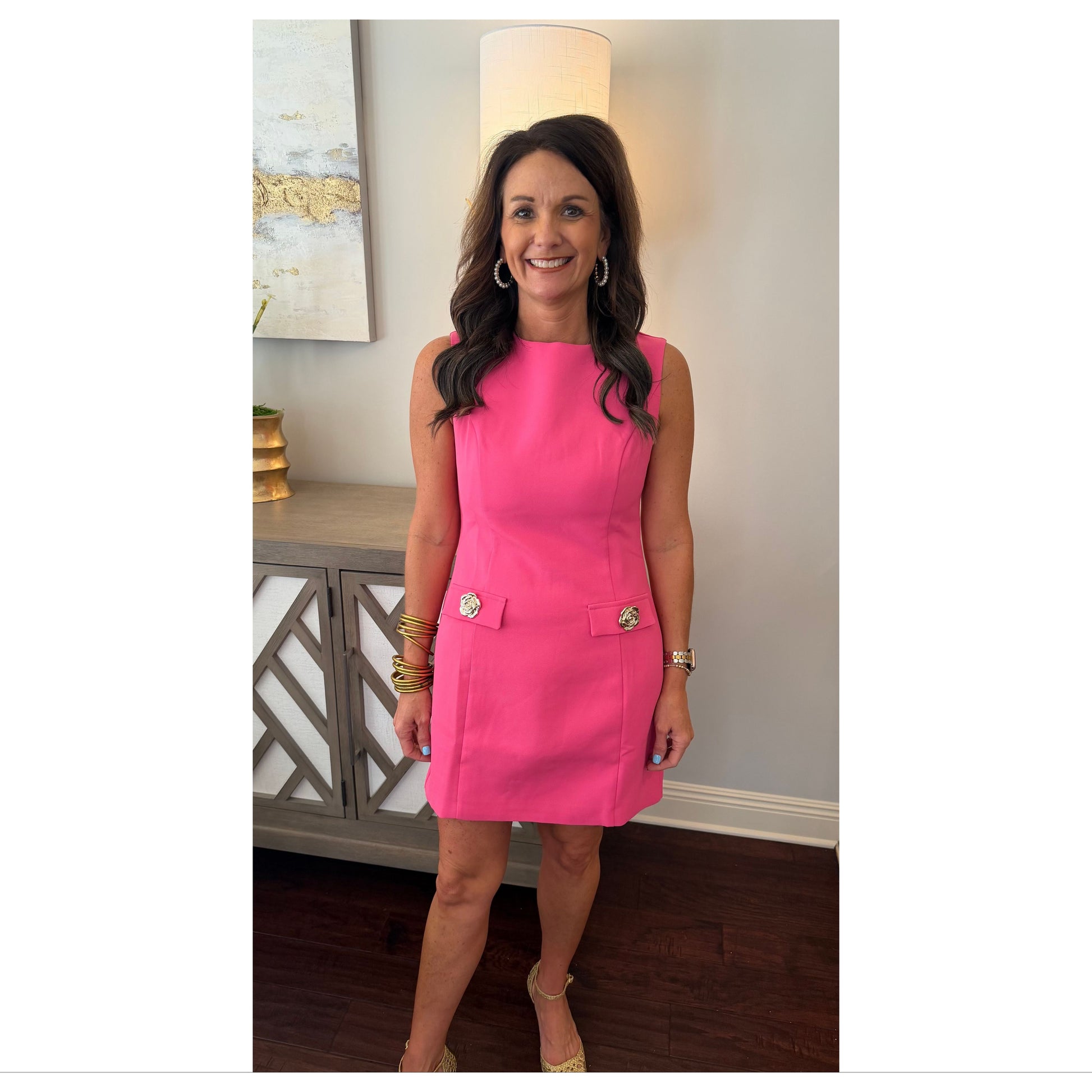 Woman wearing a pink dress standing in a room with a lamp and decorative items.