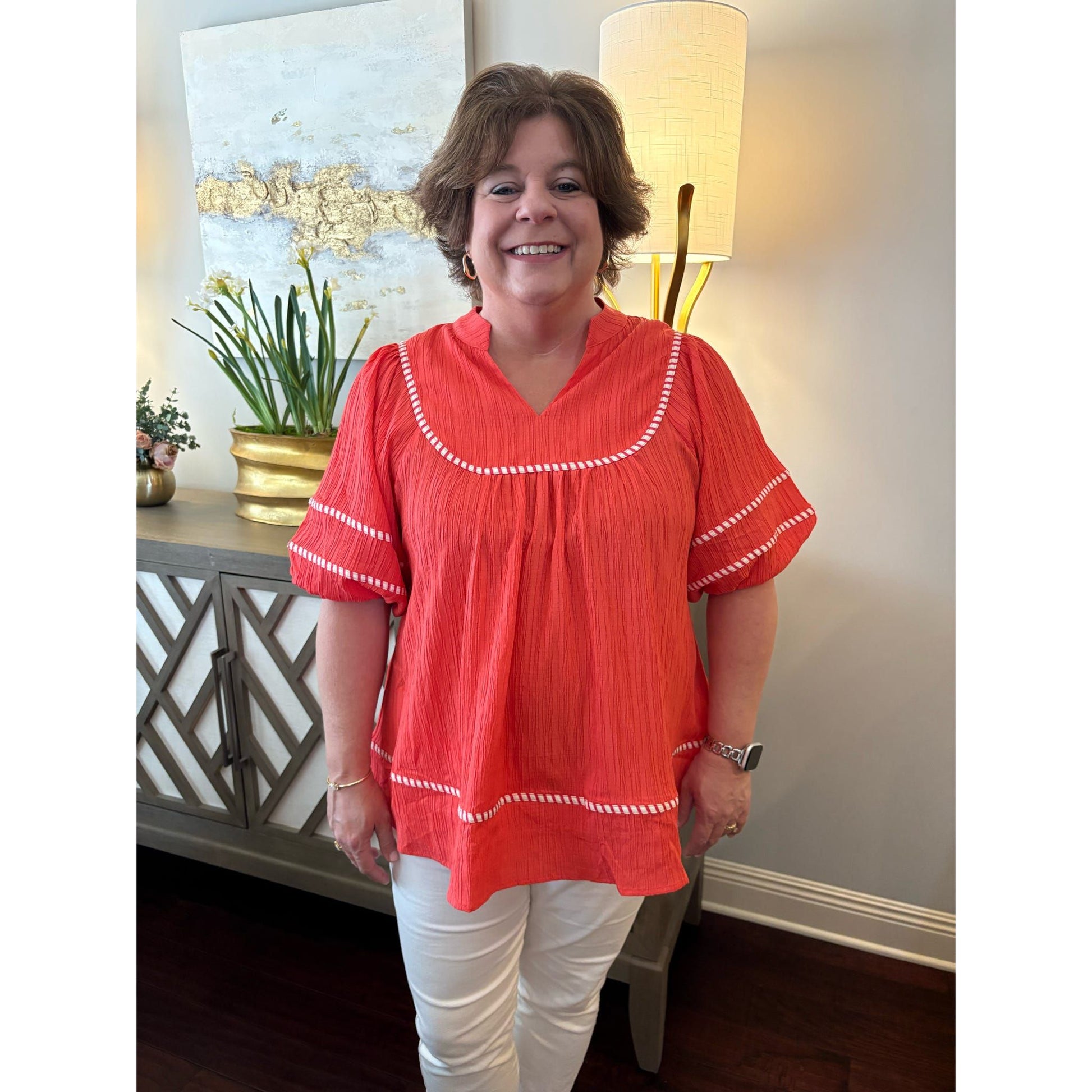 Woman wearing a red blouse with white trim in a room with a lamp and decorative items.