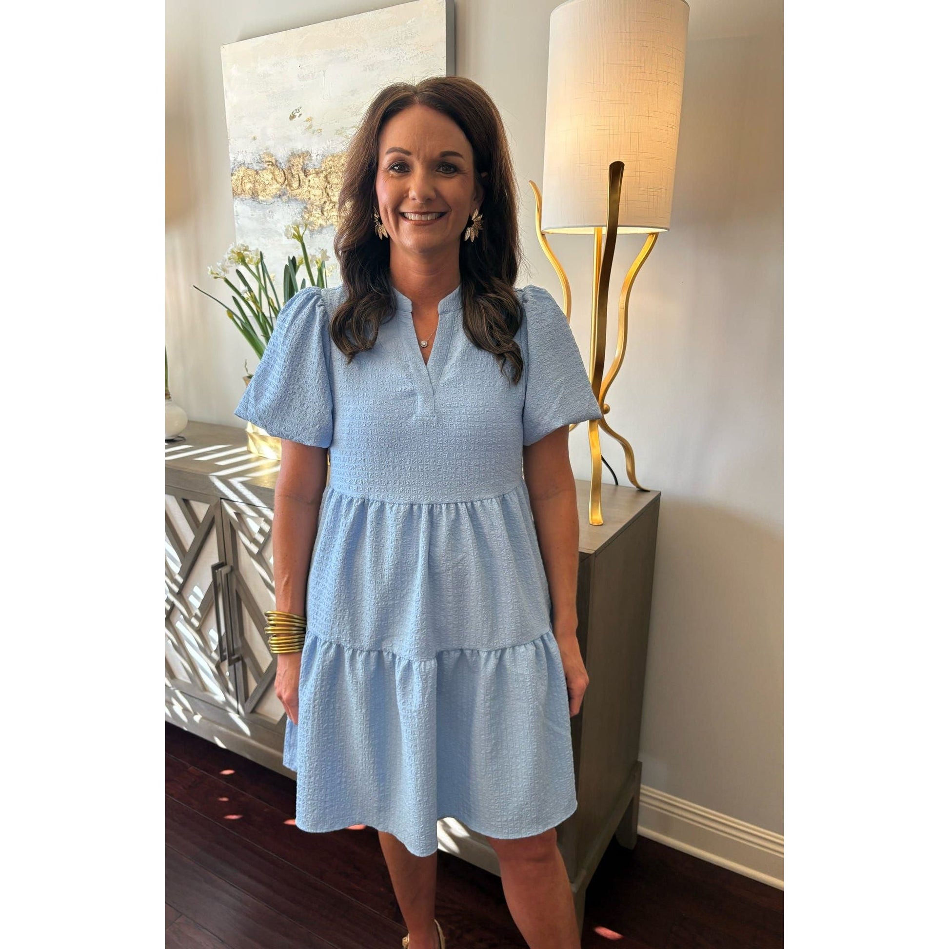 Woman in a light blue dress standing in an elegant room with lamps and artwork.