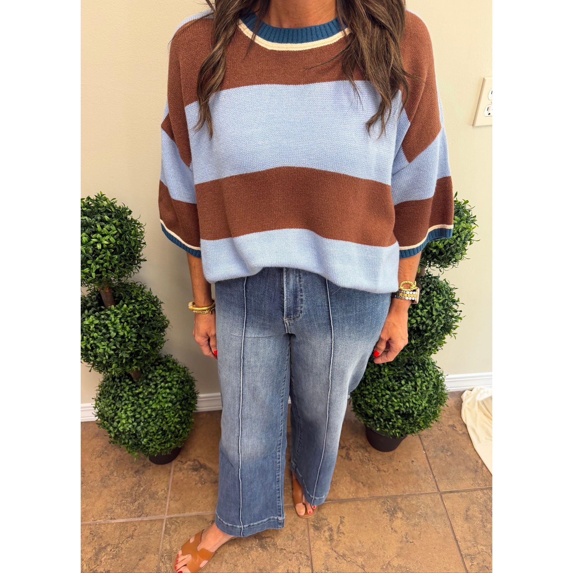 A woman wearing a brown and light blue striped knit sweater with blue jeans, standing indoors between two potted green plants.