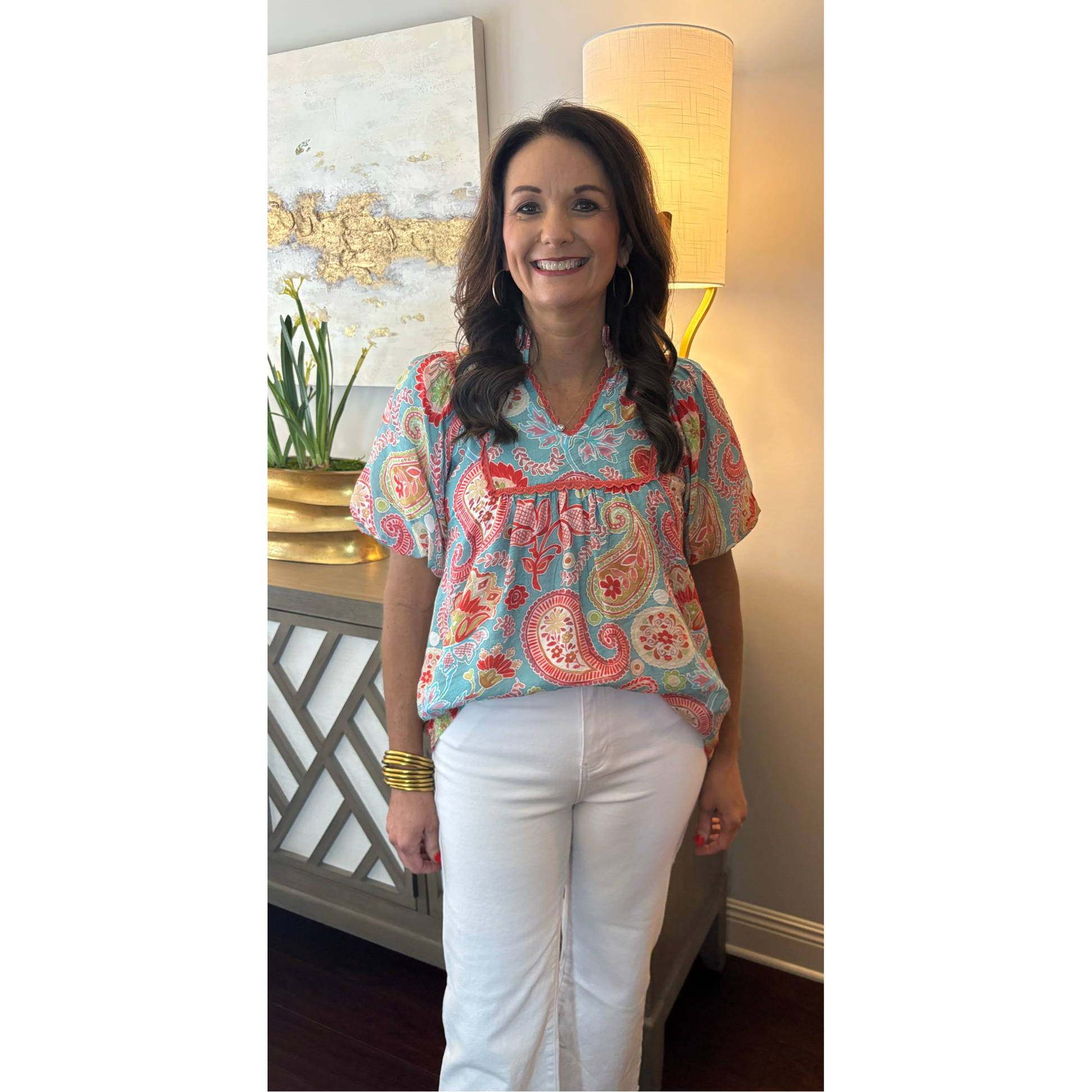 Woman wearing a colorful paisley blouse and white pants indoors.