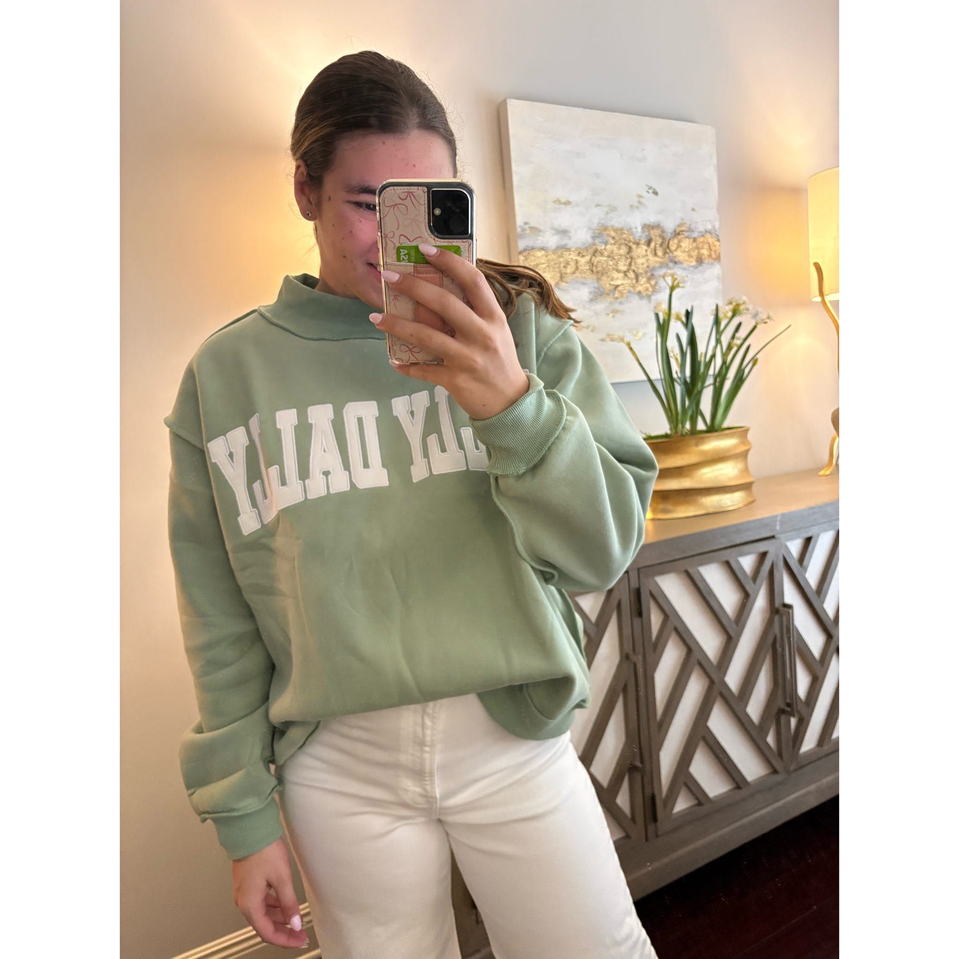 Person wearing a green sweatshirt with text, taking a mirror selfie in a room.