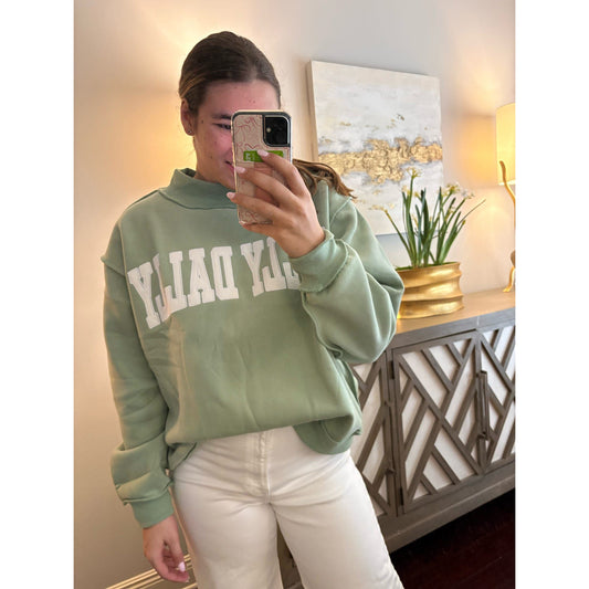 Person wearing a green sweatshirt with text, taking a mirror selfie in a room.