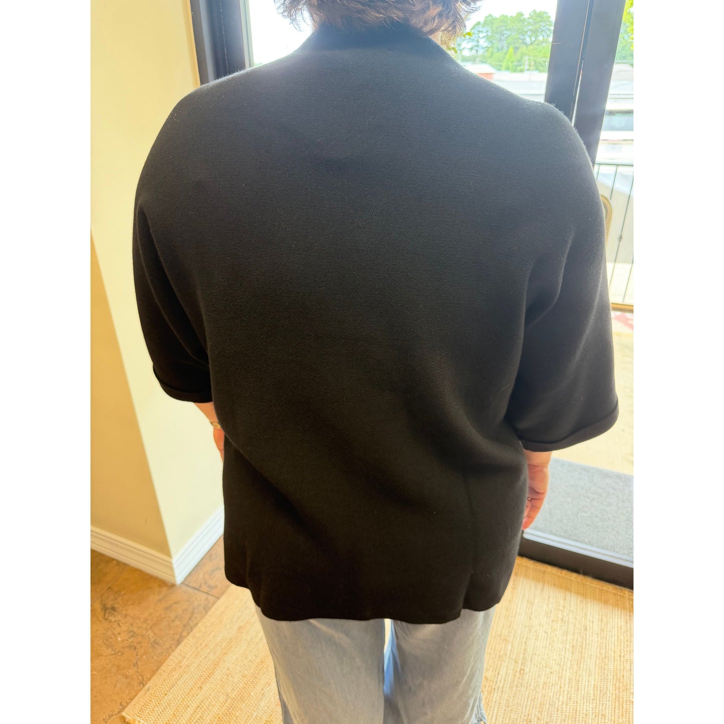 A person viewed from behind wearing a black short-sleeve knit top or cardigan, standing indoors near a glass door and light wood floor.