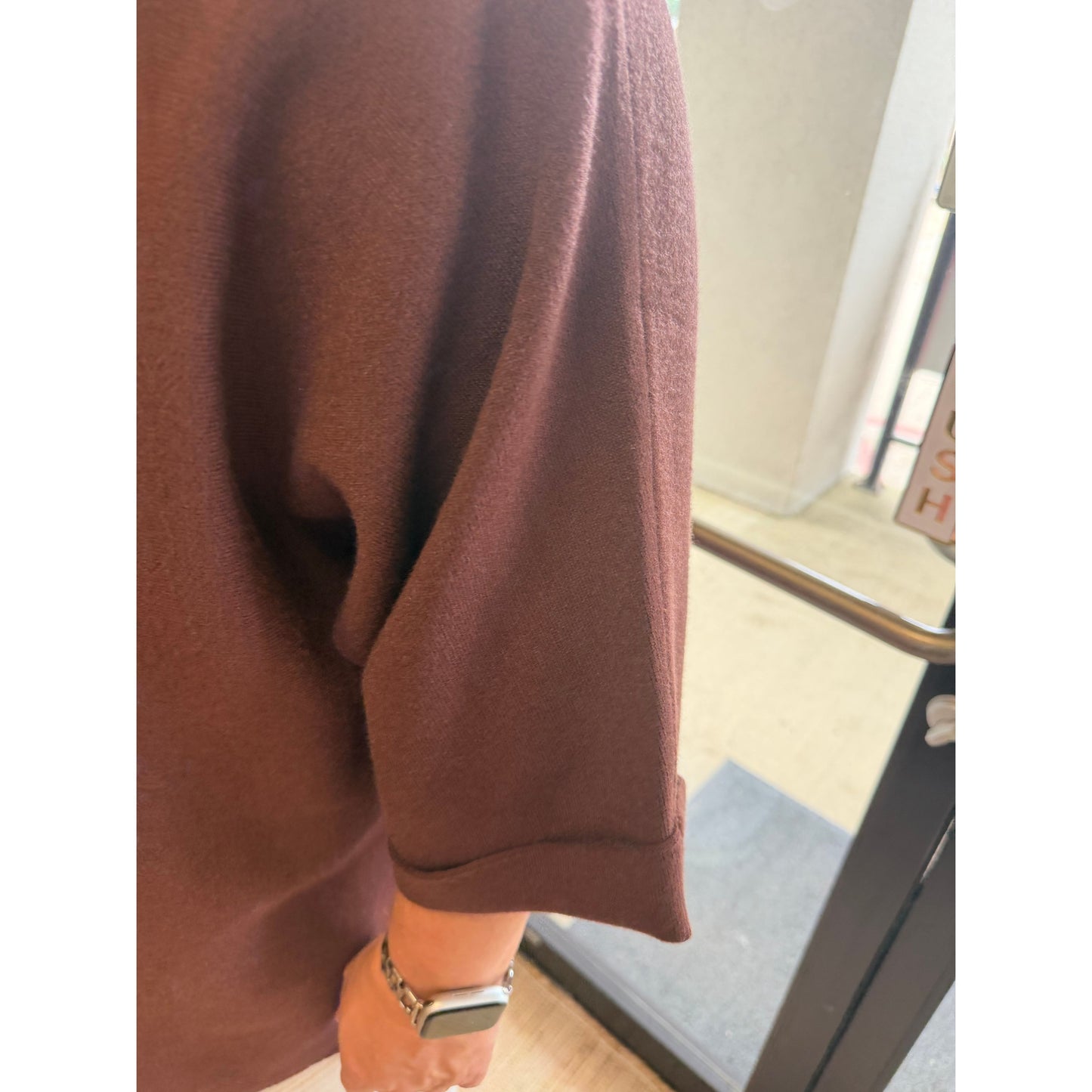 A close-up of a person wearing an oversized brown knit top or sweater, showing the sleeve and cuff, with a watch on the wrist and a real indoor doorway in the background.