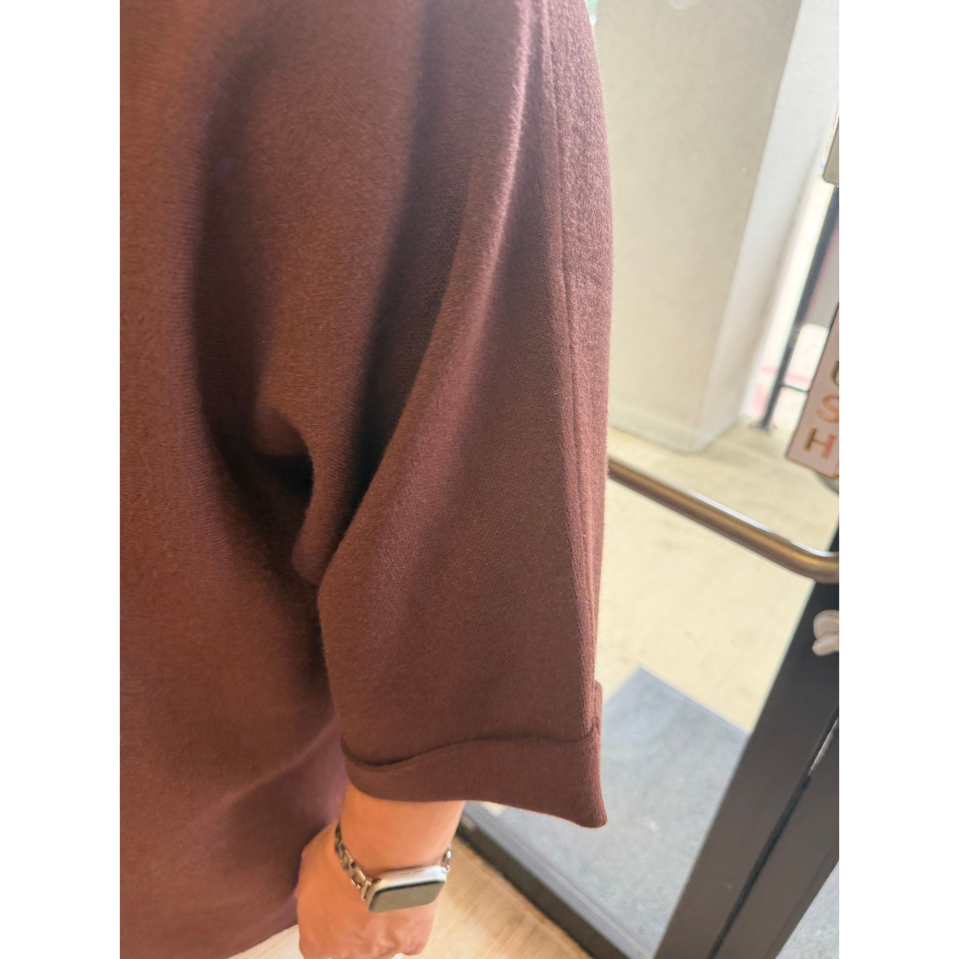 A close-up of a person wearing an oversized brown knit top or sweater, showing the sleeve and cuff, with a watch on the wrist and a real indoor doorway in the background.