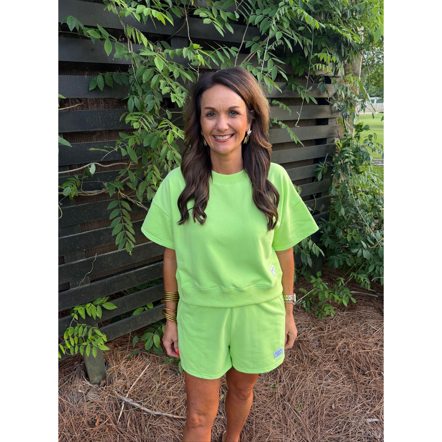 Woman wearing a bright green outfit standing in front of a wooden wall with plants.
