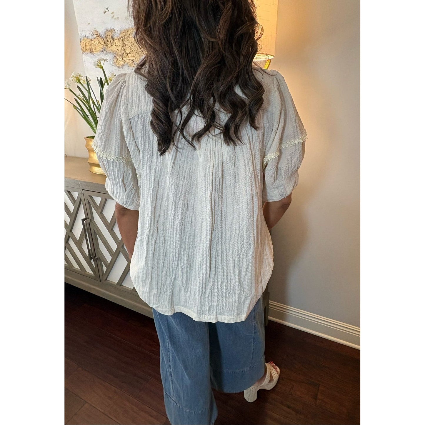 Person wearing a light-colored blouse and blue jeans standing in a room with a decorative wall piece.