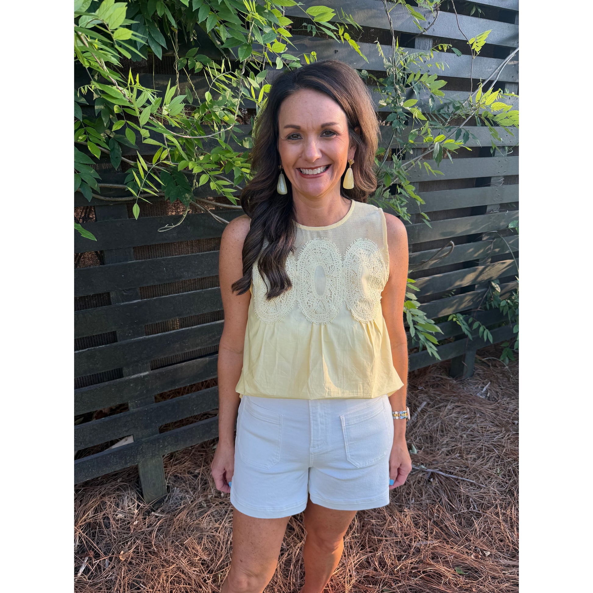 Woman wearing a light yellow sleeveless top and white shorts standing in front of a wooden fence with greenery.