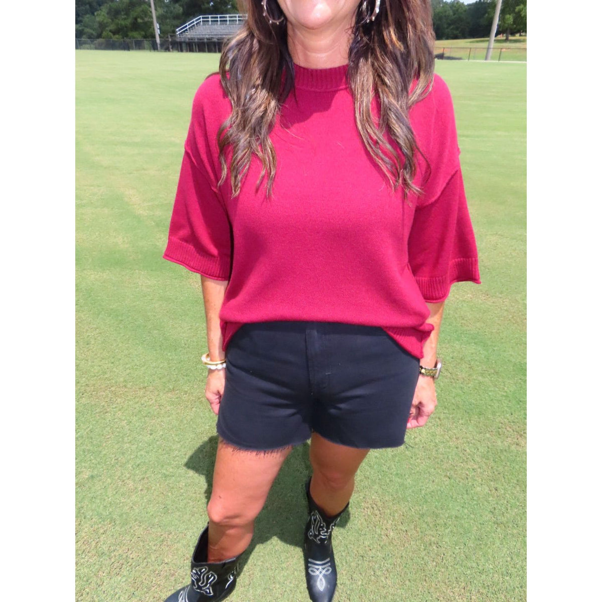 A woman wearing a bright pinkish-red short-sleeve knit top, black shorts, and black cowboy boots standing on a green grass field.